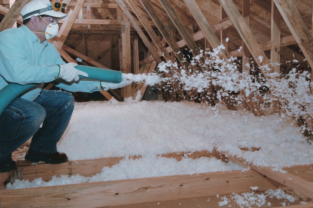 Technician crouching in an unfinished attic, wearing face mask, blowing white loose-fill insulation onto the attic floor.